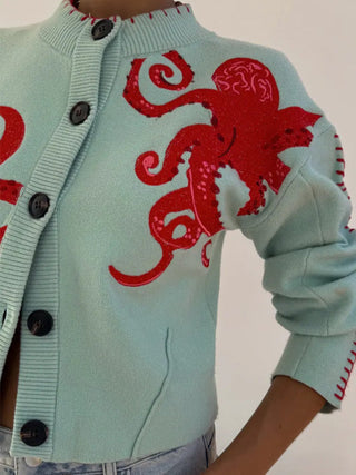 Cardigan bleu clair manches longues, motif pieuvre rouge sur l’épaule, boutons noirs visibles.