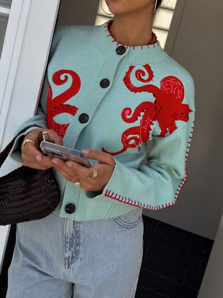 Cardigan en tricot bleu clair, motif pieuvre rouge sur le devant, boutons noirs visibles.